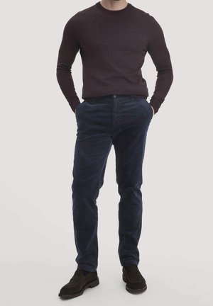 Homme portant une chemise à manches longues marron foncé, un pantalon en velours côtelé bleu marine et des chaussures marron foncé, les mains dans les poches, debout sur un fond uni.