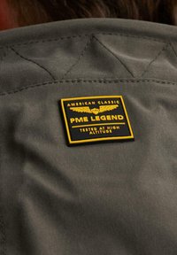 Tessuto grigio con una finitura testurizzata presenta un patch nero e giallo che reca la scritta "PME LEGEND" con ali e "TESTATO AD ALTE ALTITUDINI."