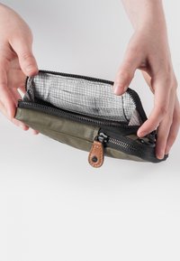 Olive green nylon pouch with black zipper, tan leather accent, and grid-patterned interior; held open to show inside compartments.