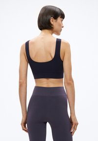 Moteris su trumpais tamsiais plaukais, vilkinti juodą sportinį liemenėlę ir tamsias aukšto liemens leginsus, stovinti nugara į žiūrovą ant paprasto fono.