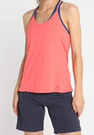 Kobieta ubrana w koralowy, bezrękawnik sportowy na granatowym biustonoszu sportowym i granatowe krótkie spodenki do kolan na białym tle.