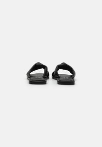 Sandales noires au design épuré, dotées de deux brides torsadées, de semelles plates et d'une texture lisse. Apparence minimaliste et moderne.