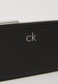 Portafoglio in pelle nera con superficie testurizzata, hardware della cerniera in argento e logo "ck" impresso sul davanti. Caratterizzato da un design semplice ed elegante.