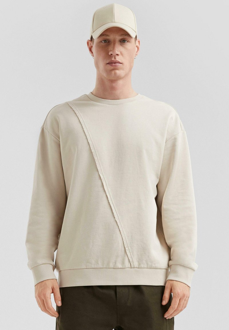 Beige sweatshirt met ronde hals en een diagonaal gestikte panelen en ribgebreide boorden. Zachte, gestructureerde stof. Gecombineerd met een bijpassende pet.