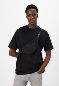 Black T-shirt with loose fit, paired with a black crossbody bag featuring a zipper and adjustable strap. Model stands casually with hands in pockets.