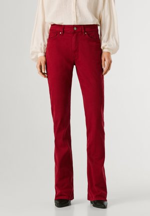 Personne portant un pantalon rouge à jambe droite associé à une chemise blanche ample à manches longues et des chaussures noires, debout devant un arrière-plan uni.