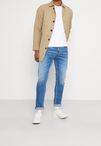 En knäppt jacka över en vit t-shirt, i kombination med ljusblå jeans och vita sneakers. Slätt tyg och figursydd design.