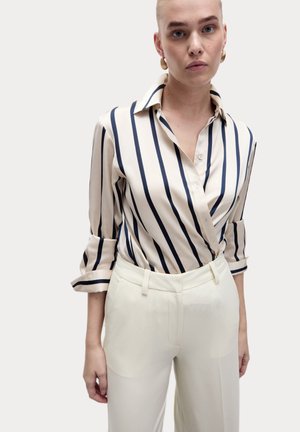 Crème blouse met donkerblauwe verticale strepen, kraag en knoopsluiting; gecombineerd met wit hoge taille broek. Glad stoftextuur.