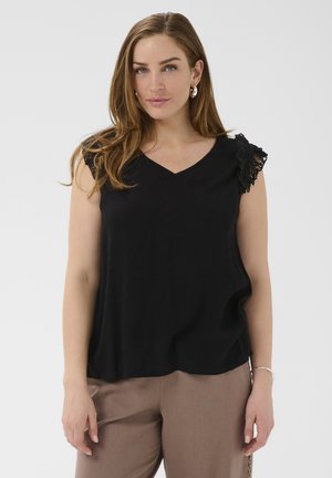 Femme aux longs cheveux bruns portant un haut noir sans manches avec des épaules en dentelle et un pantalon beige, debout devant un fond uni.