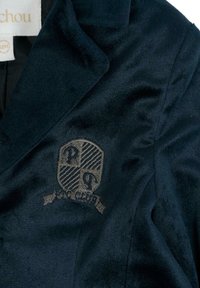 Granatowy aksamitny blezer z usztywnianym kołnierzem i szarą wyszywaną tarczą. Gładka tekstura i dopasowany krój z wyraźnymi liniami.