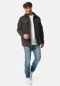 Heren donkergrijze corduroy jas met een zachte zwarte bontkraag, voorzakken en een rits, gecombineerd met lichtblauwe versleten jeans en witte sneakers.