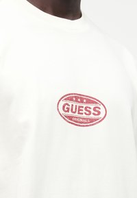 Camiseta blanca de algodón con un logo ovalado rojo que presenta el texto "GUESS ORIGINALS" en el centro, cuello redondo con textura acanalada.