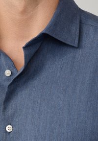 Donkerblauw, gestructureerd overhemd met een visgraatpatroon; heeft een kraag en twee knopen zichtbaar bij de hals.