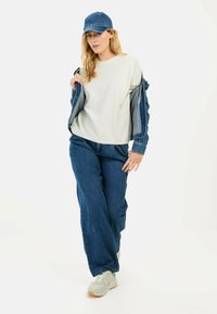 Denimjack over een lichtgrijze textuurtop, gecombineerd met loszittende blauwe jeans en beige sneakers. De pet is ook van denim, wat de look compleet maakt.