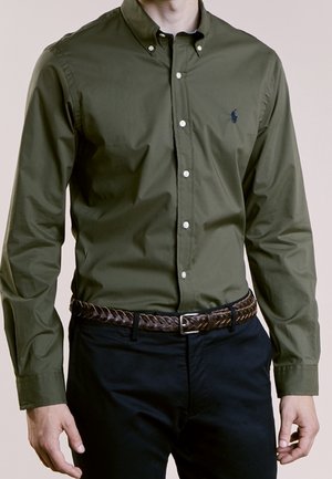 Olijfgroene button-up shirt met een klassieke kraag, witte knopen en een klein logo op de borst, gecombineerd met een gevlochten bruine riem en donkere broek.