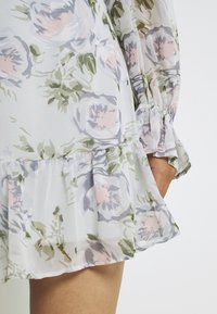 Robe florale en tissu transparent à motifs de roses pastel et de feuilles vertes. L'ourlet froncé et les poignets froncés rehaussent le design.