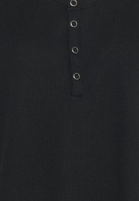 Svart våffelstickad tyg med fyra metalltryckknappar vertikalt placerade på en placket.