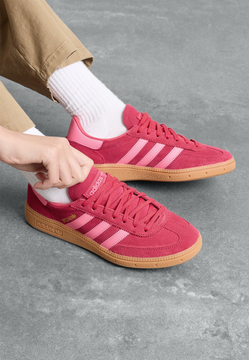 Sapatos Adidas em camurça rosa vibrante com riscas rosa mais claras, bico redondo e sola de borracha texturizada. Meias brancas às riscas complementam o visual.