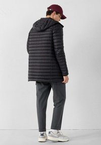 Giacca a vento nera con cuciture orizzontali e cappuccio, abbinata a pantaloni grigi e sneakers bianche, vista da dietro.
