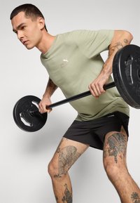 Lichtgroen sport t-shirt met korte mouwen, gecombineerd met zwarte shorts. Een verzwaarde halter wordt vastgehouden tijdens een oefening, met gedetailleerde been tatoeages.