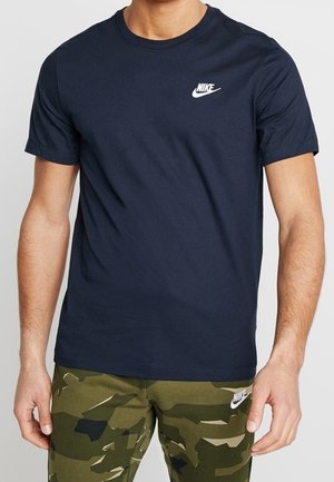 Man wearing a navy blue Nike t-shirt and green camouflage Nike shorts, standing with arms relaxed at sides.