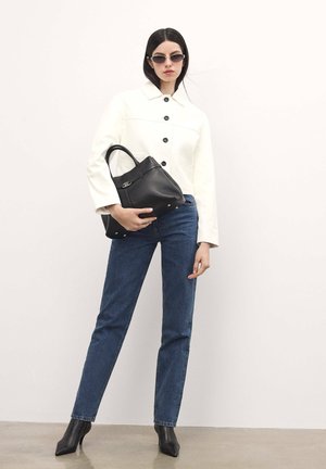 White cropped jacket with black buttons, blue denim jeans, and black heeled ankle boots. Black handbag held in the right hand.