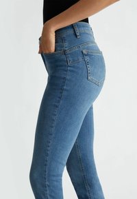 Jeans ajustados de denim azul con una cintura alta, que cuentan con bolsillos laterales y traseros, y un diseño clásico de cinco bolsillos. Textura suave.