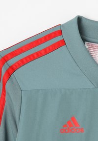 Szary sportowy t-shirt z czerwonymi trzema paskami na ramionach, żebrowana tekstura oraz małe czerwone logo Adidas z przodu.