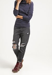 Marinsvett med hög krage och präglad design, kombinerat med slitna svarta jeans med hål, och vinröda snörstövlar.