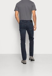 Mörkblå jeans med slim fit, tillverkade av mjukt bomullstyg, bakfickor och en svart märkesetikett på midjebandet. Kombinerat med grå skor.