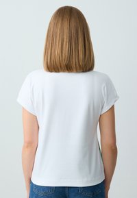 Weißes Kurzarmoberteil mit lässigem Schnitt und glatter Textur. Der Rücken zeigt einen abgerundeten Saum und kurze, gerollte Ärmel, kombiniert mit blauen Jeans.