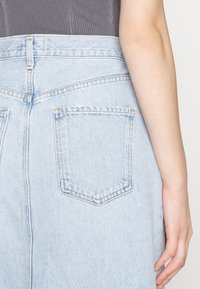 Personne portant un jean en denim bleu clair et un haut côtelé gris foncé, visible de la taille jusqu'au milieu de la cuisse avec le bras droit visible.