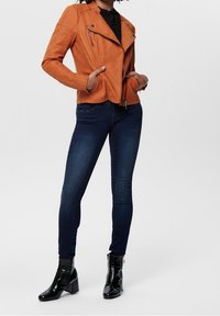 Pomarańczowa skórzana kurtka motocyklowa z zamkami błyskawicznymi i wykończeniem na ramionach, zestawiona z ciemnoniebieskimi obcisłymi dżinsami i błyszczącymi czarnymi kozakami do kostek.