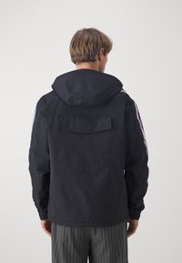 Mężczyzna w czarnej kurtce z kapturem z paskowymi detalami na rękawach i szarych spodniach w cienkie prążki, zwrócony tyłem na gładkim tle.