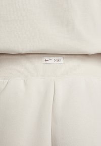 Pantalones deportivos beige claro con cintura acanalada. La etiqueta de la marca presenta el logo de Nike y el nombre en marrón claro sobre un fondo blanco.