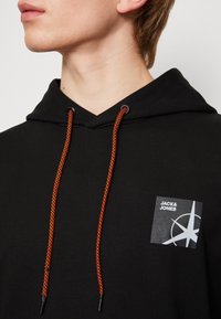 Černá mikina s kapucí a oranžovými šňůrkami, která má na přední části nášivku s logem a designem kompasu. Vyrobena z měkké látky, má uvolněný střih.