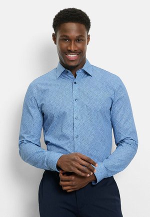 Lächelnder Mann, der ein hellblaues, gemustertes Hemd mit langen Ärmeln und dunkle Hosen trägt, vor einem weißen Hintergrund stehend.