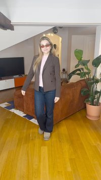 Femme portant des lunettes de soleil, un blazer, un jean et des bottes, debout sur un sol en bois dans un salon avec un canapé marron et une grande plante en pot.