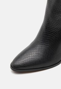 Black ankle boot with a textured, hexagonal pattern. Pointed toe and smooth finish enhance its sleek silhouette. Black sole.