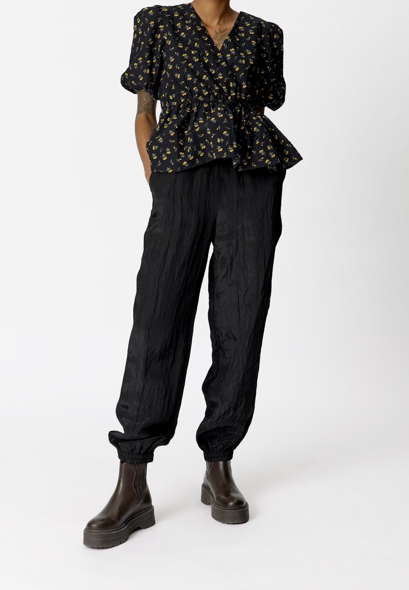 Top peplum nero a fiori con maniche a sbuffo, abbinato a pantaloni neri svasati e texturizzati. Modella indossa stivaletti marroni chunky alla caviglia.