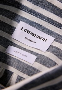 Etiqueta de camisa de lino de algodón con el texto "LINDBERGH", "AJUSTE RELAJADO" y "ALGODÓN LINO" sobre un fondo a rayas azul y blanco.