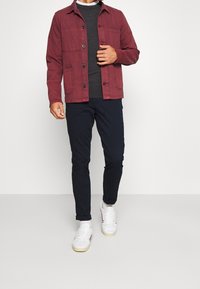 Man som bär uppvikt mörka byxor, vita sneakers med strumpor, mörkgrå tröja och knäppt röd jacka mot en neutral bakgrund.