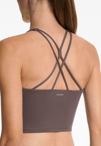 Reggiseno sportivo marrone con spalline incrociate e logo OYSHO. Il materiale è liscio con una superficie testurizzata, design aderente.