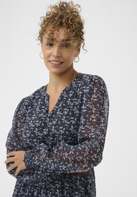 Femeie cu părul buclat, purtând cercei circulari din aur și o bluză lungă cu mâneci din material transparent și imprimeu floral închis, zâmbind cu brațele încrucișate.