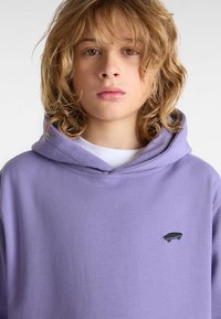 Lila Kapuzenpullover aus weichem Material mit einer Kängurutasche und einem kleinen schwarzen Skateboard-Logo. Die Kapuze hat eine abgerundete Form und verstärkte Nähte.