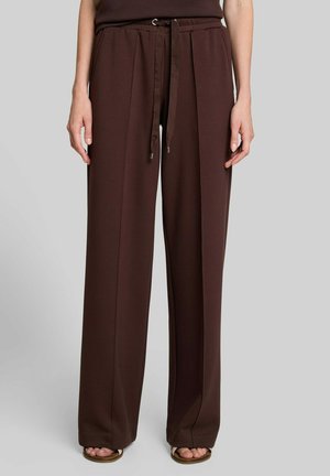 Mujer con pantalones marrones de tiro alto y pierna ancha con cordón delantero, combinados con sandalias de punta abierta sobre un fondo gris liso.