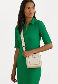 Μπεζ τσάντα ώμου με υφή, χρυσά εξαρτήματα και έντονο λογότυπο. Φορεμένη με ένα εφαρμοστό πράσινο ριπ φόρεμα.