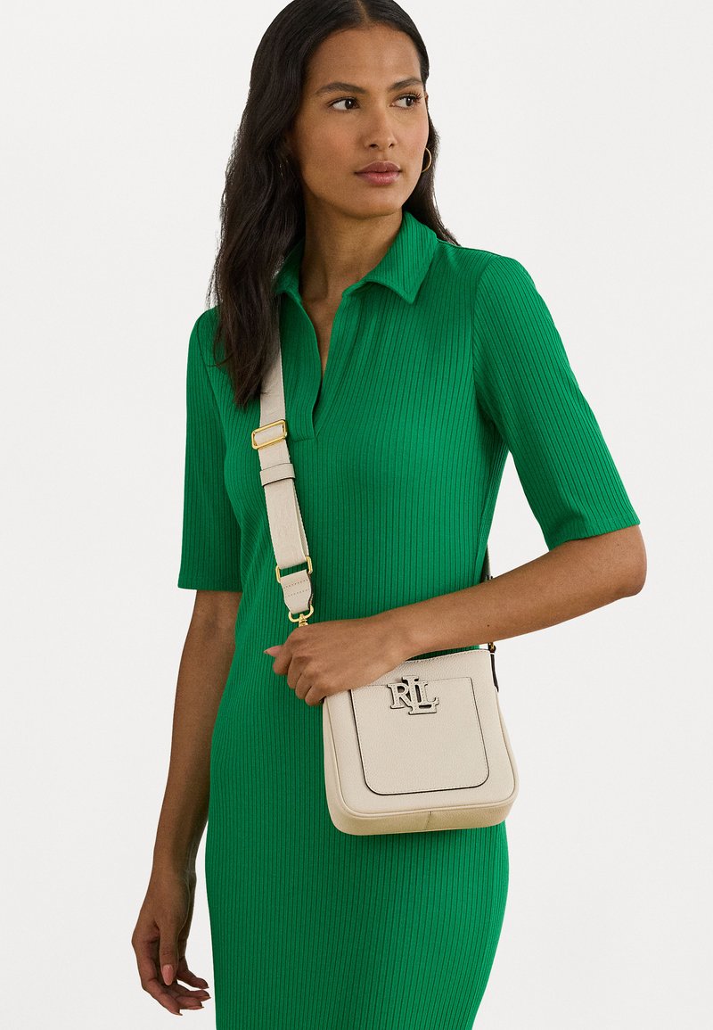 Μπεζ τσάντα ώμου με υφή, χρυσά εξαρτήματα και έντονο λογότυπο. Φορεμένη με ένα εφαρμοστό πράσινο ριπ φόρεμα.