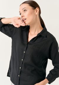 Camicia nera in lino con bottoni, caratterizzata da colletto, maniche lunghe e fessure laterali, con bottoni chiari a contrasto e una finitura testurizzata.