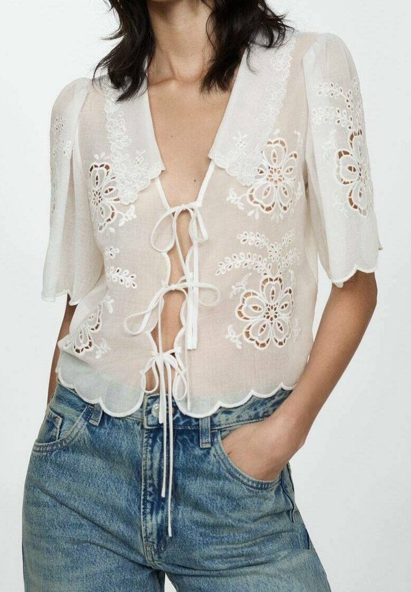 Femme portant un chemisier blanc transparent avec broderie florale, bords festonnés, manches courtes, noué devant, et un jean bleu en denim avec la main dans la poche.